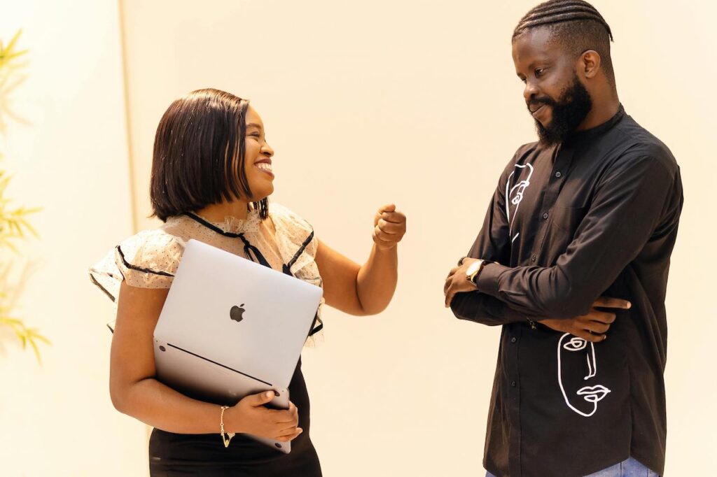 A woman holding a laptop speaks with a colleague while reviewing CAC registration documents for a new business.